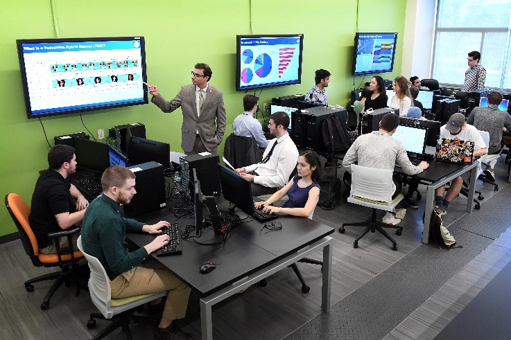 students at a table in a classroom looking a screens with a professor instructing