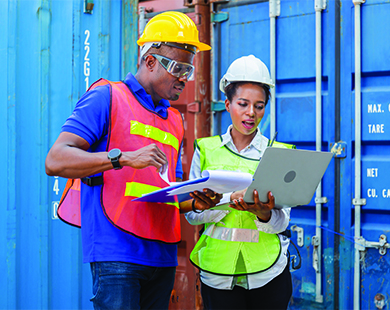 two people in hard hats and vests at a construction site looking at a laptop together